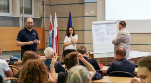 Spotkanie konsultacyjne ws. hałasu ulicznego w Urzędzie Miasta Gdyni — funkcjonariusz policji, moderatorka przy flipcharcie z postulatami mieszkańców i przedstawiciel urzędu, w tle uczestnicy spotkania