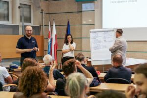 Spotkanie konsultacyjne ws. hałasu ulicznego w Urzędzie Miasta Gdyni — funkcjonariusz policji, moderatorka przy flipcharcie z postulatami mieszkańców i przedstawiciel urzędu, w tle uczestnicy spotkania