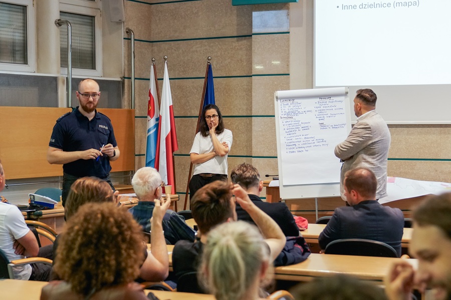 Spotkanie konsultacyjne ws. hałasu ulicznego w Urzędzie Miasta Gdyni — funkcjonariusz policji, moderatorka przy flipcharcie z postulatami mieszkańców i przedstawiciel urzędu, w tle uczestnicy spotkania