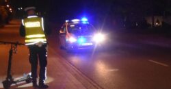 Policjant przy zabezpieczonej hulajnodze elektrycznej na miejscu nocnego wypadku drogowego, w tle radiowóz z włączonymi sygnałami świetlnymi
