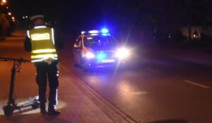 Policjant przy zabezpieczonej hulajnodze elektrycznej na miejscu nocnego wypadku drogowego, w tle radiowóz z włączonymi sygnałami świetlnymi
