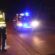 Policjant przy zabezpieczonej hulajnodze elektrycznej na miejscu nocnego wypadku drogowego, w tle radiowóz z włączonymi sygnałami świetlnymi